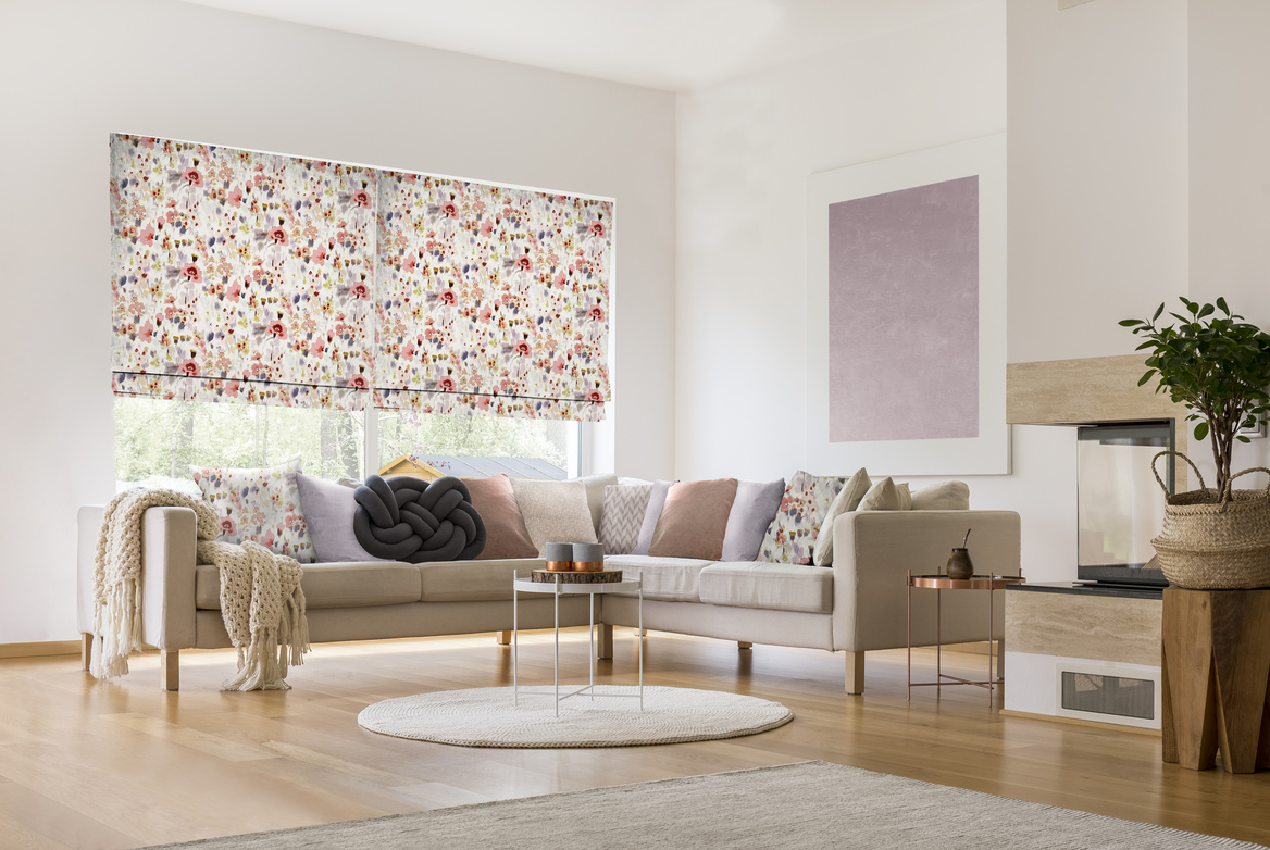 Neutral lounge with L shaped sofa with accent cushions in soft pink and floral matching two Roman blinds at window behind with medium sized pink and beige flowers, accent grey knot-shaped cushion on sofa.