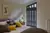 Bedroom with ivory walls, bed dressed in ivory and chocolate brown bedding and an angular shaped tall window dressed in black shaped shutters closed, with louvres open.