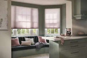 Contemporary kitchen with banquette seating under bay windows dressed in soft pink Duette® blinds drawn halfway, co-ordinating cushions on blue seating, kitchen counters in soft grey with white top.