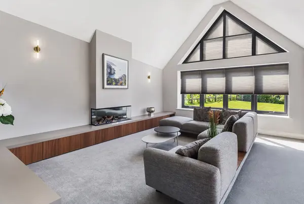 Pale grey lounge with gable end windows and doors dressed in beige duette blinds, closed on top, lowered halfway on bottom, two sofas in L shape opposite feature fireplace.