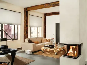 Loft apartment style lounge room with beams and feature pillar fire, lit. and two large windows dressed in pale Duette blinds slightly lowered,