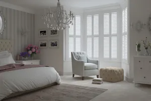 White classic-style bedroom with bay window dressed in white shutters, louvres bed on left with white bedding and dusky pink throw, white upholstered chair in bay window with pale ochre upholstered footstool, and chandelier.