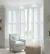Living room in pale grey with white full height shutters in a bay window, partially open, louvres open, pale grey chair in recess, pale beige pouffee in front of chair.