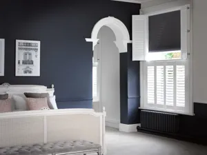 Bedroom with window dressed in white tier on tier shutters, closed with louvres open, dark blue walls, white bed with grey and dusky pink cushions, grey upholstered stool at bed, archway with white moulding.
