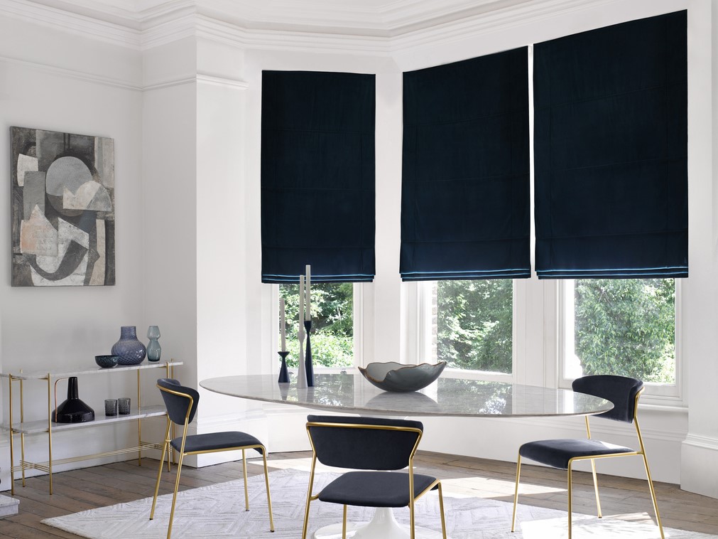Modern dining room with bay window dressed in three navy blue velvet Roman blinds, oval glass table, metallic gold chairs with blue upholstery, metallic gold side table with blue glassware.