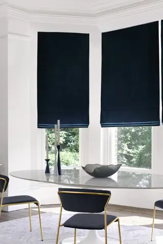 Modern dining room with bay window dressed in three navy blue velvet Roman blinds, oval glass table, metallic gold chairs with blue upholstery, metallic gold side table with blue glassware.