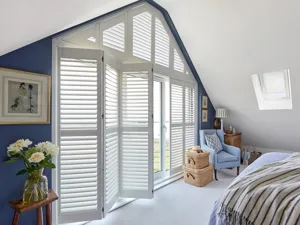 Gable end azure blue wall in bedroom with other walls in white, gable end window and doors dressed in white shutters, louvres open and tracked door shutters partially tracked open, shaed shutter above closed, white carpet, pale blue upholstered chair in right corner, small table with white roses in left, bed with blue and white bedding.