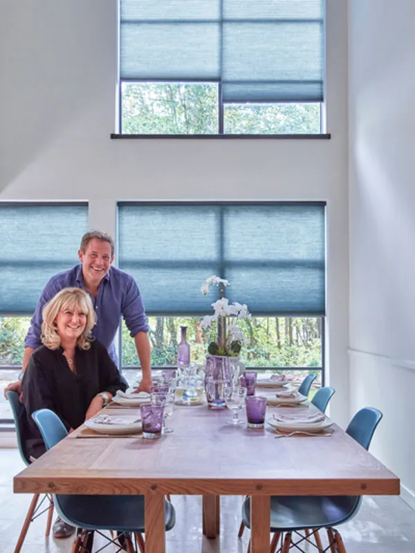 Matt and Lisa Tebbutt in dining room with double height windows dressed in mid blue duette blinds, wooden dining table and blue chairs.