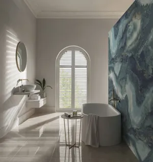 white arched shutters open letting light into a modern white bathroom with a green and blue patterned feature wall behind the bath