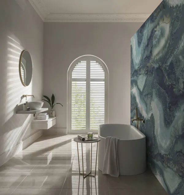 white arched shutters open letting light into a modern white bathroom with a green and blue patterned feature wall behind the bath