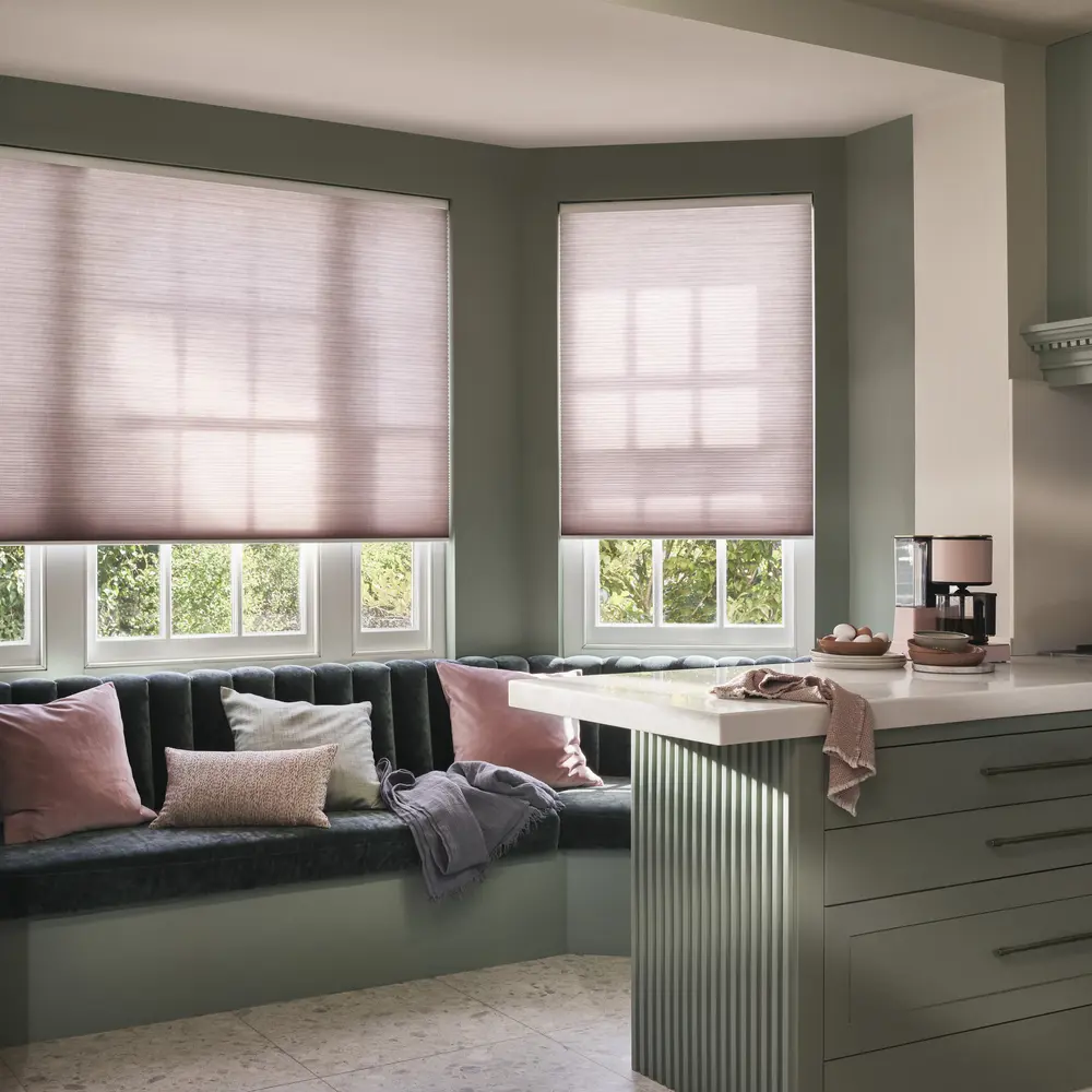 Contemporary kitchen with banquette seating under bay windows dressed in soft pink Duette blinds drawn halfway, co-ordinating cushions on blue seating, kitchen counters in soft grey with white top.