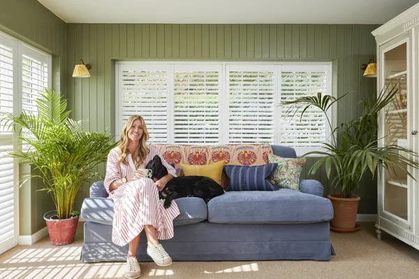Someone sat on a grey sofa in a living room with bright green walls and plants pots, while the windows are fitted with tier on tier white shutters