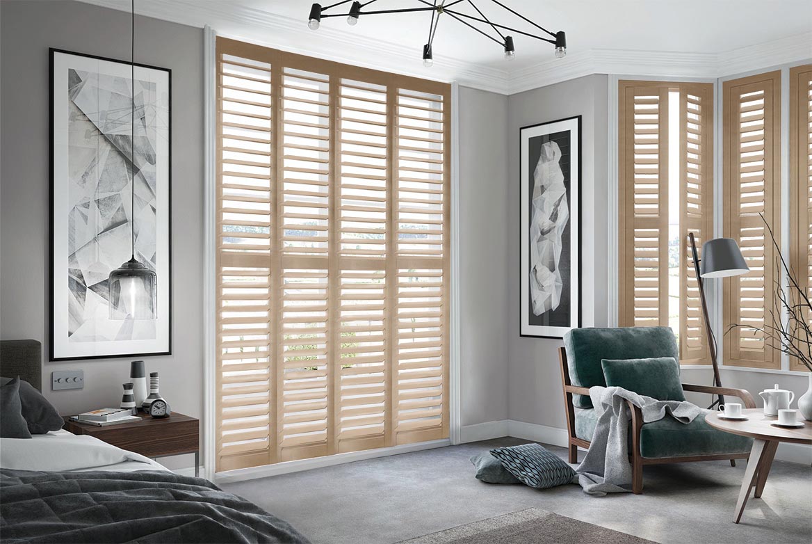 Roasted Rye Shutters arranged in a tier formation are fitted to tall windows in a bedroom decorated with a neutral grey colour. The room has a bed, side table, armchair and coffee table