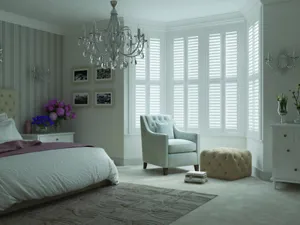 White classic-style bedroom with bed on right with white bedding and dusky pink throw, white upholstered chair in bay window with pale ochre upholstered footstool, chandelier and bay window dressed in white shutters, louvres open.
