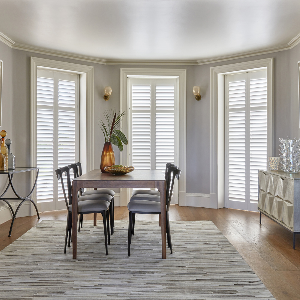 Dining room with wood floor, rug in grey tones, three floor to ceiling windows in an extended bay dressed in white shutters, closed with louvres open, wooden table and chairs with grey cushioned seats, amber vase and greenery on table.
