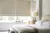 Tight shot of neutral bedroom, bed with white bedding, grey/brown throw across bed, small cream wrap on top, pale wood beside table with cream lamp and large windows dressed in two Roman blinds in ivory faux velvet.
