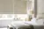 Tight shot of neutral bedroom, bed with white bedding, grey/brown throw across bed, small cream wrap on top, pale wood beside table with cream lamp and large windows dressed in two Roman blinds in ivory faux velvet.