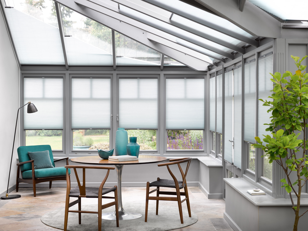Pale grey conservatory with side windows and roof dressed in pale grey blinds, three quarters lowered on sides and varying heights on roof, stone floor, grey rug under circular wooden table and two chairs with marine blue vases, marine blue low chair in top left corner with silver anglepoise lamp.