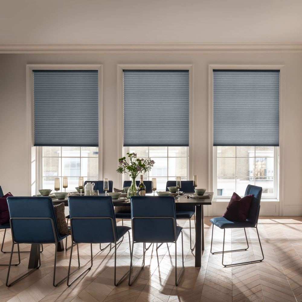 Dining room with three tall windows dressed in mid blue duette blinds, three quarters lowered, dining chairs in blue around table.