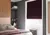 Bedroom with pink and grey bedding, pink headboard against mid-wood wall, grey wall to side with white inset with silver radiator and white sash window dressed with plum chenille Roman blind lowered three quarters.