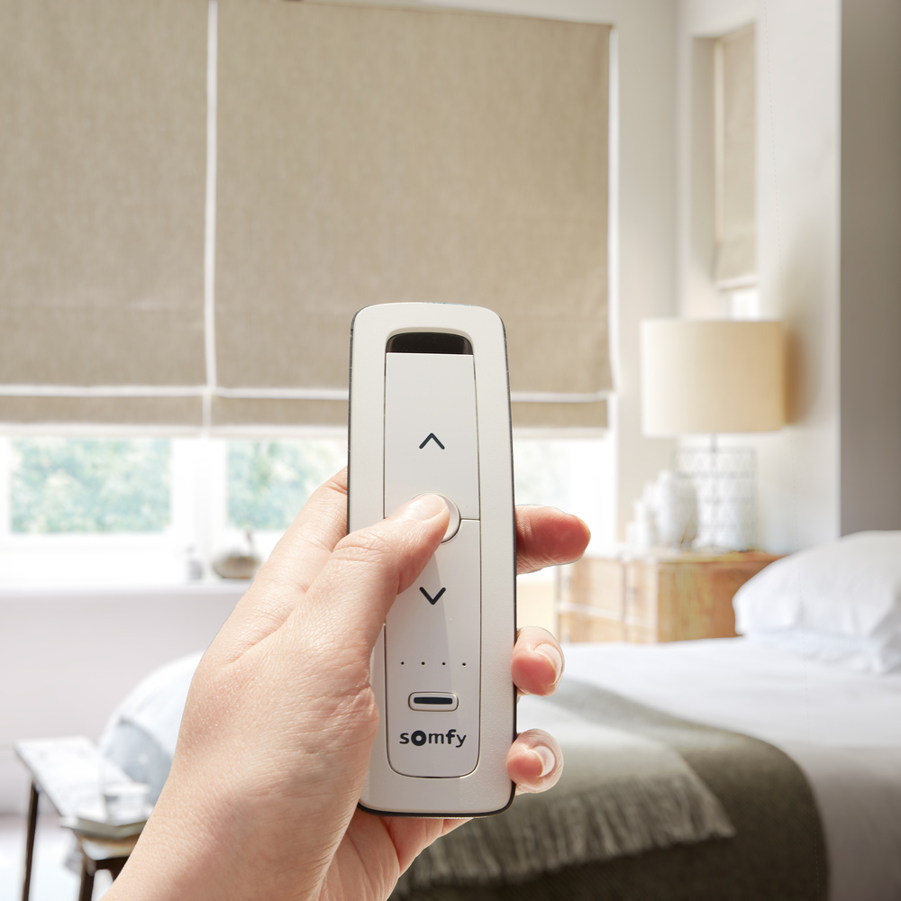 beige electric roman blind in a bedroom with a hand holding a somfy remote infront of it