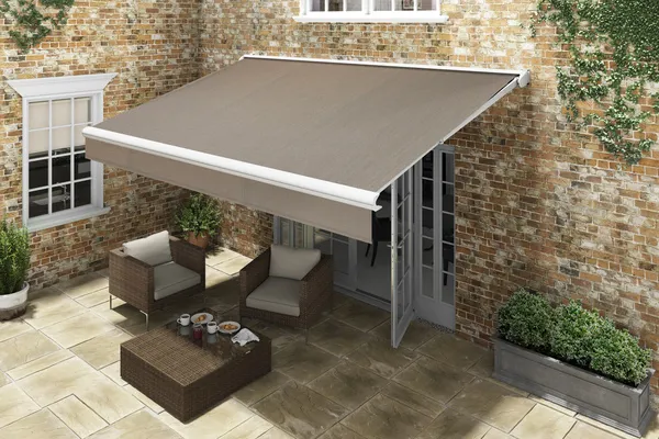 Exterior of traditional pale brown stone house with a mid brown awning extended over the flagged patio with dark brown raffia chairs with cream cushions and low table. 