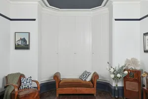White room with wooden chair, wooden piano stool in front of a bay window dressed with white solid shutters, closed.