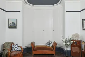 White room with wooden chair, wooden piano stool in front of a bay window dressed with white solid shutters, closed.