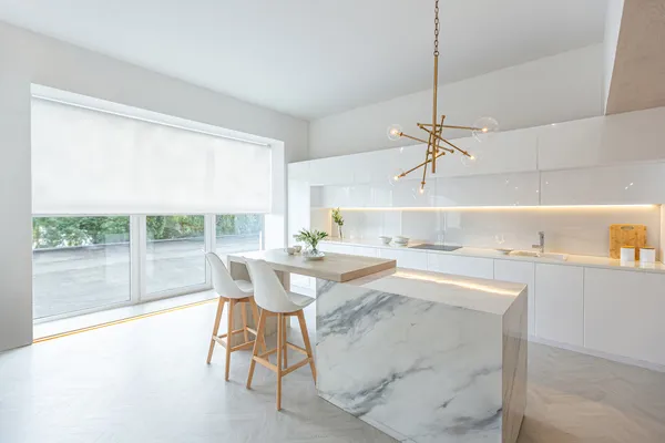 Open-plan white kitchen with trio of window and doors dressed in white roller blind with marble island with white chairs and modern pendant light.