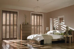 Neutral bedroom with wooden bed with white bedding and beige accessories two floor to ceiling windows dressed in mid-colour wood shutters, louvres open with light rays filling room.