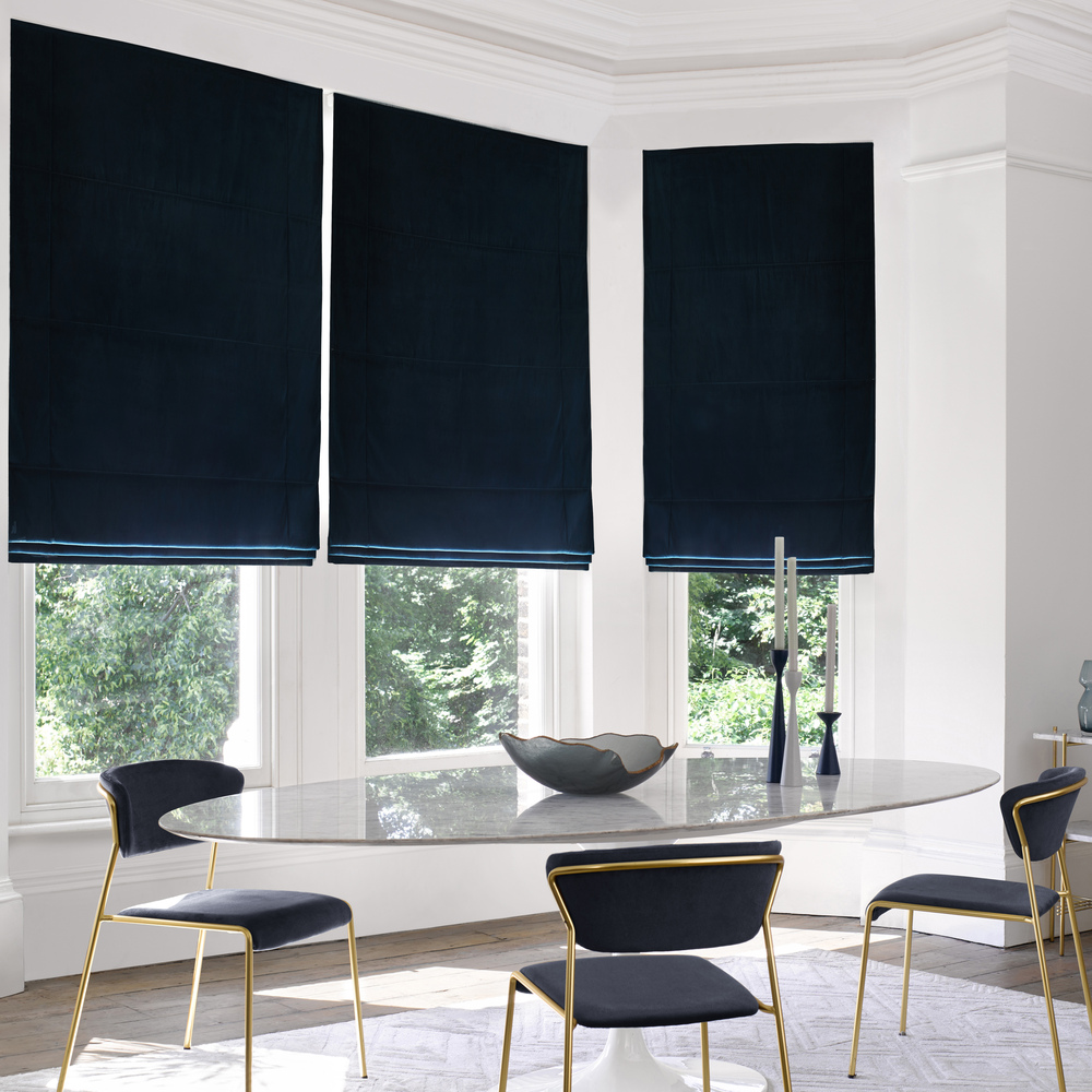 Modern dining room with bay window dressed in three navy blue velvet Roman blinds, oval glass table, metallic gold chairs with blue upholstery, metallic gold side table with blue glassware.