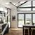 Large kitchen with gable end windows and doors dressed in soft white duette blinds at varying heights, parquet floor, dark grey units and kitchen island with sink and wooden stools.
