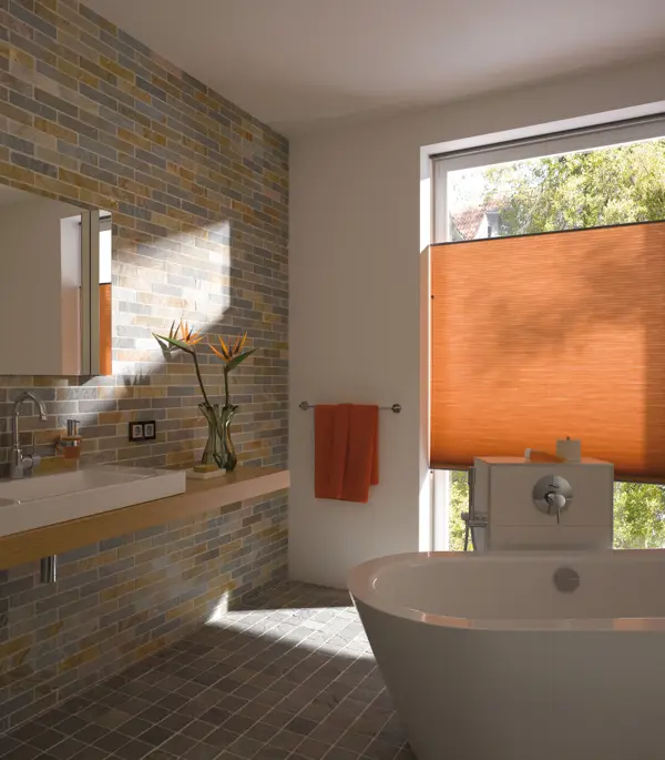 Bathroom with picture window in white wall, dressed in a cadmium orange blind in the middle of the window, side wall and floor tiled in metro stile in shades of brown, grey and copper, white bathtub in centre, pale wood shelf with white sink, cadmium orange towel on rail by window.