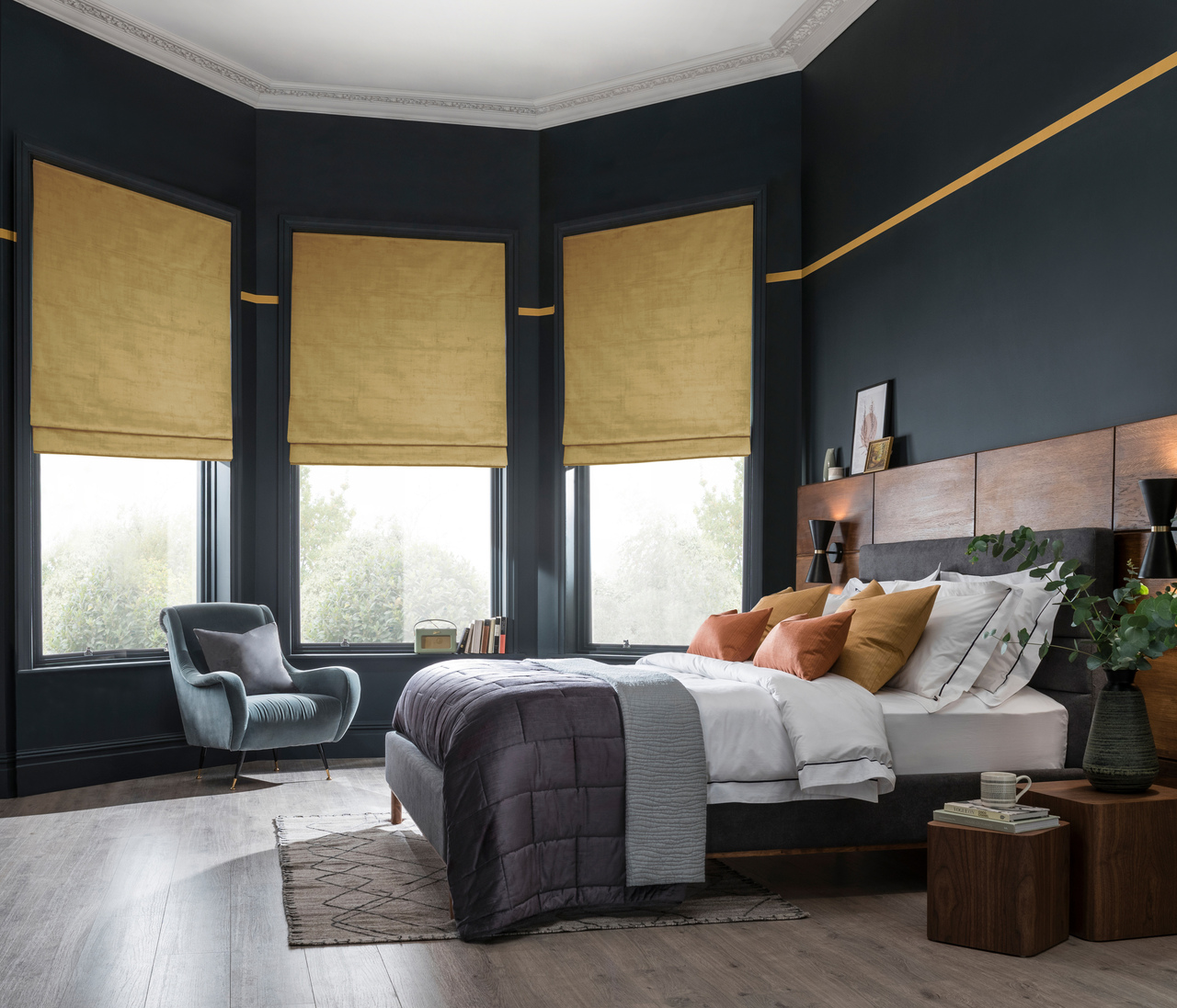 Large bedroom with wooden floor, bay window with three ochre faux velvet Roman blinds, navy walls, bed with white bedding and grey quilted throw,. 