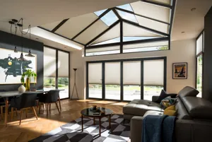 Open plan living diner with wood floor, brown leather sofa, modern table and chairs, angled glass ceiling, gable end windows and doors and additional doors all dressed in pale brown-grey Duette blinds at varying levels.