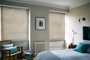 Pale grey bedroom with bed dressed in pale blue bedding, wooden chair in corner with blue cushions and two windows dressed in pale stone-grey coloured wood venetian blinds, lowered, louvres tilted.