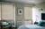Pale grey bedroom with bed dressed in pale blue bedding, wooden chair in corner with blue cushions and two windows dressed in pale stone-grey coloured wood venetian blinds, lowered, louvres tilted.