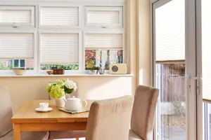 Conservatory with magnolia wall, table small dining table and suede chairs with doors and windows with cream pleated blinds at varying heights.