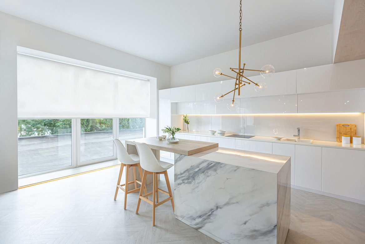 Open-plan white kitchen with trio of window and doors dressed in white roller blind with marble island with white chairs and modern pendant light.