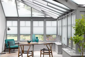 Pale grey conservatory with stone floor, grey rug under circular wooden table and two chairs with marine blue vases, marine blue low chair in top left corner with silver anglepoise lamp and side windows and roof dressed in pale grey blinds, three quarters lowered on sides and varying heights on roof.