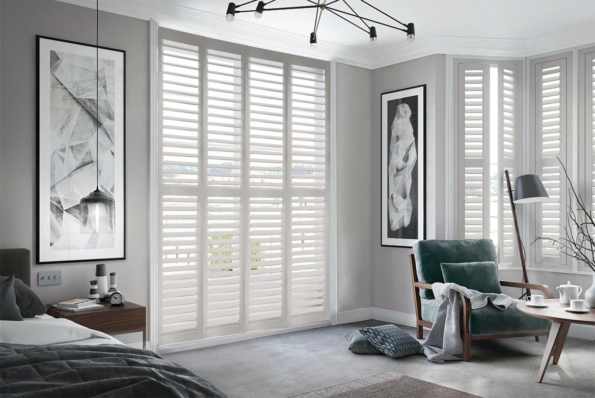 True Grey Shutters have been fitted to tall windows in a bedroom. The Shutters are Grey coloured while the bedroom is decorated with grey tones.