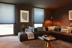 Burnt ochre living room with corner brown velvet sofa, modern copper-based table in centre and two large windows dressed in deep blue duette blinds three-quarters lowered.