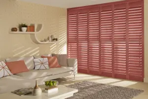 Living room in cream with brown rug, pale beige sofa with terracotta cushions in plain and patterned fabric and floor to ceiling shutters in terracotta, louvres open.