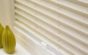 Close up of a pale peach pleated blind, lowered with a darker toned veining pattern, with two small olive ripple-textured vases on the window sill.