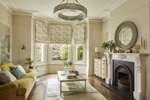 Living room in neutral tones with wood floor, chartreuse sofa, round mirror above ornate mantlepiece, round pendant light and bay window dressed in Roman blind with a leaf pattern in greens and browns on a cream background, lowered halfway.
